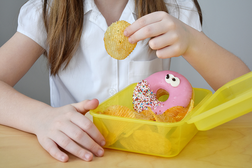 A school aged girl eating chips and a donut out of a lunchbox.
