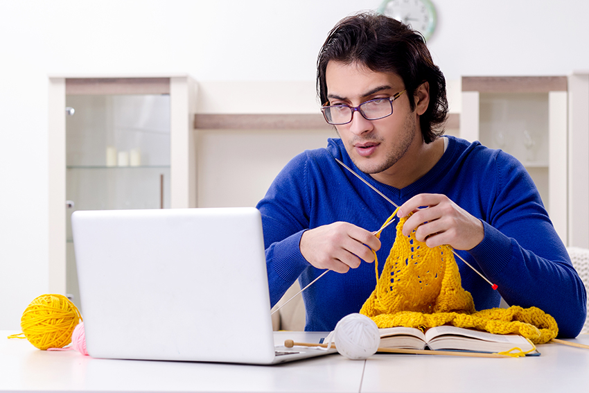 A stock image of a man knitting while looking at a laptop.